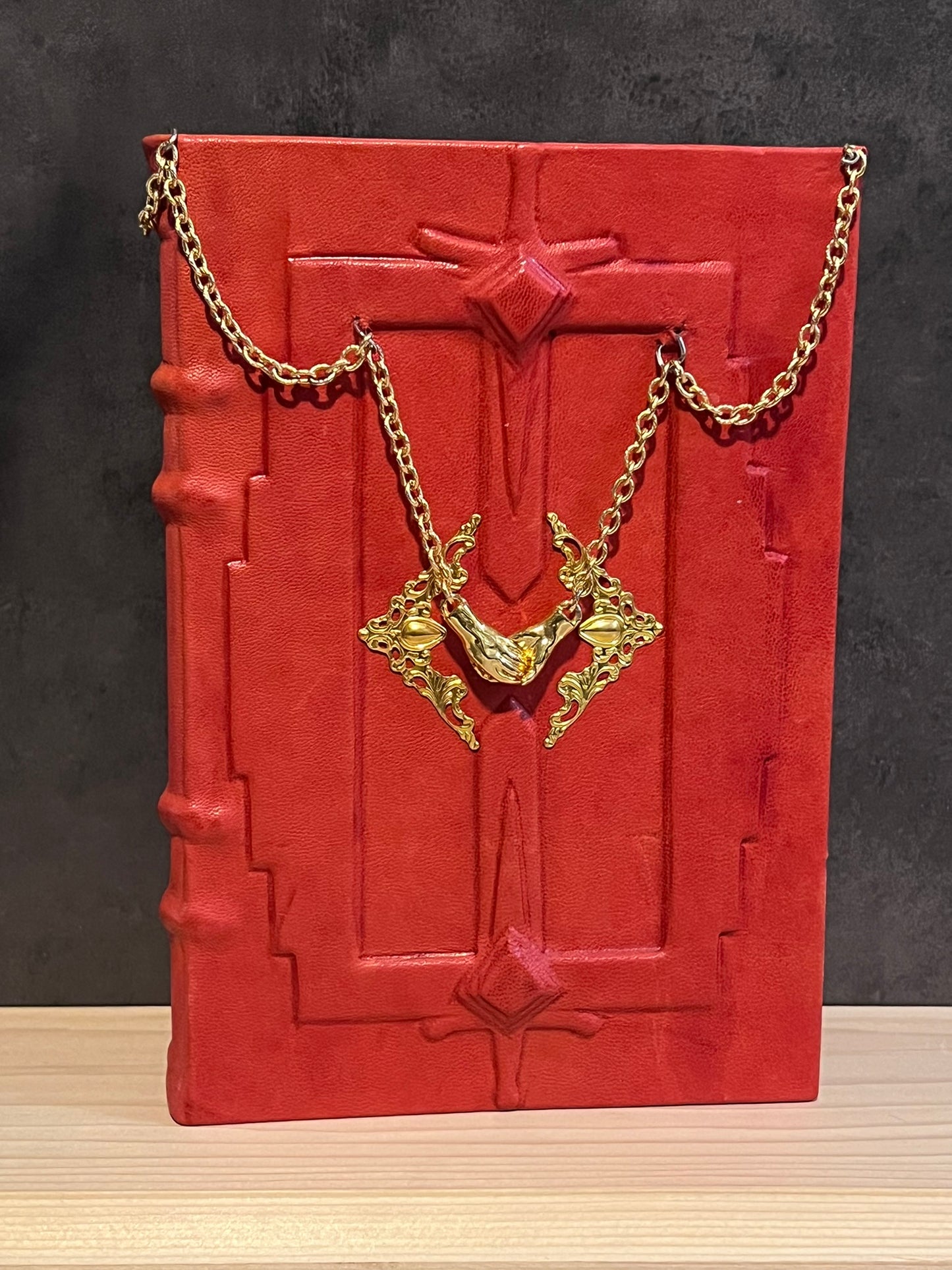 a red book with gold chains draped and leading to a pair of magnetic lapsed hands. Above and below the hands are embossed swords. To the sides of the hands are angular filigree onlays