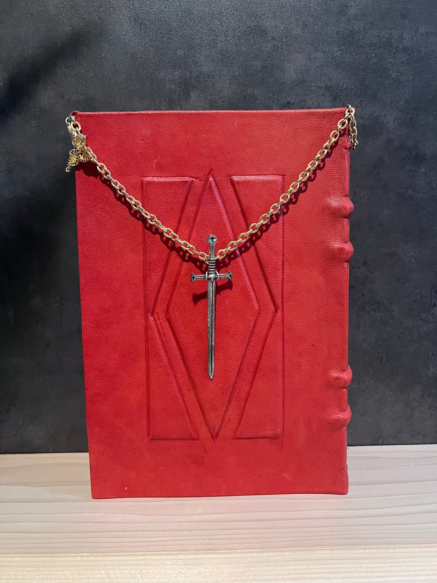 The back of the book. Also covered in red leather with a raised diamond in the center surrounded by triangles. Hanging by chain in the center is a silver sword.